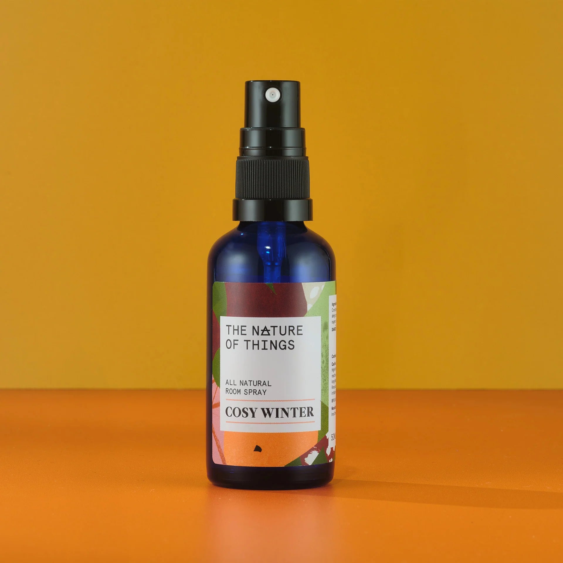 Blaue 50-ml-Sprühflasche mit schwarzem Pumpkopf und einem Etikett mit der Aufschrift „The Nature of Things All Natural Room Spray Cozy Winter“ vor orangefarbenem Hintergrund.
