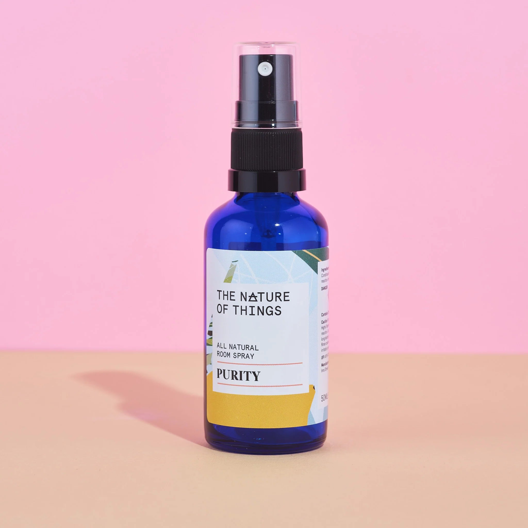 Blaue 50ml-Sprühflasche des Purity Room Sprays von The Nature of Things mit gelb-weißem Etikett und schwarzem Sprühkopf vor rosa-beigem Hintergrund.