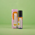Blauer 10-ml-Roll-On und dazugehörige rosa Umverpackung mit Orangen- und Gänseblümchen-Illustrationen vor einem grünen Hintergrund.