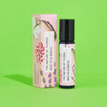 Blauer 10-ml-Roll-On und dazugehörige rosa Umverpackung mit weißen Blüten-Illustrationen vor einem grünen Hintergrund.