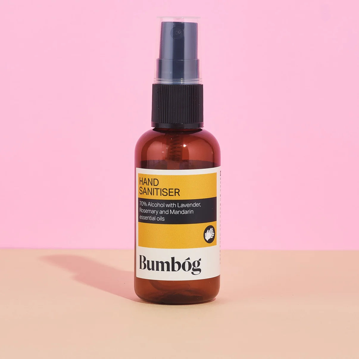 Braune 50ml Plastikflasche des Bumbóg Hand Sprays mit gelb-schwarzem Etikett und Pumpsprüher vor einem rosa und beigen Hintergrund.