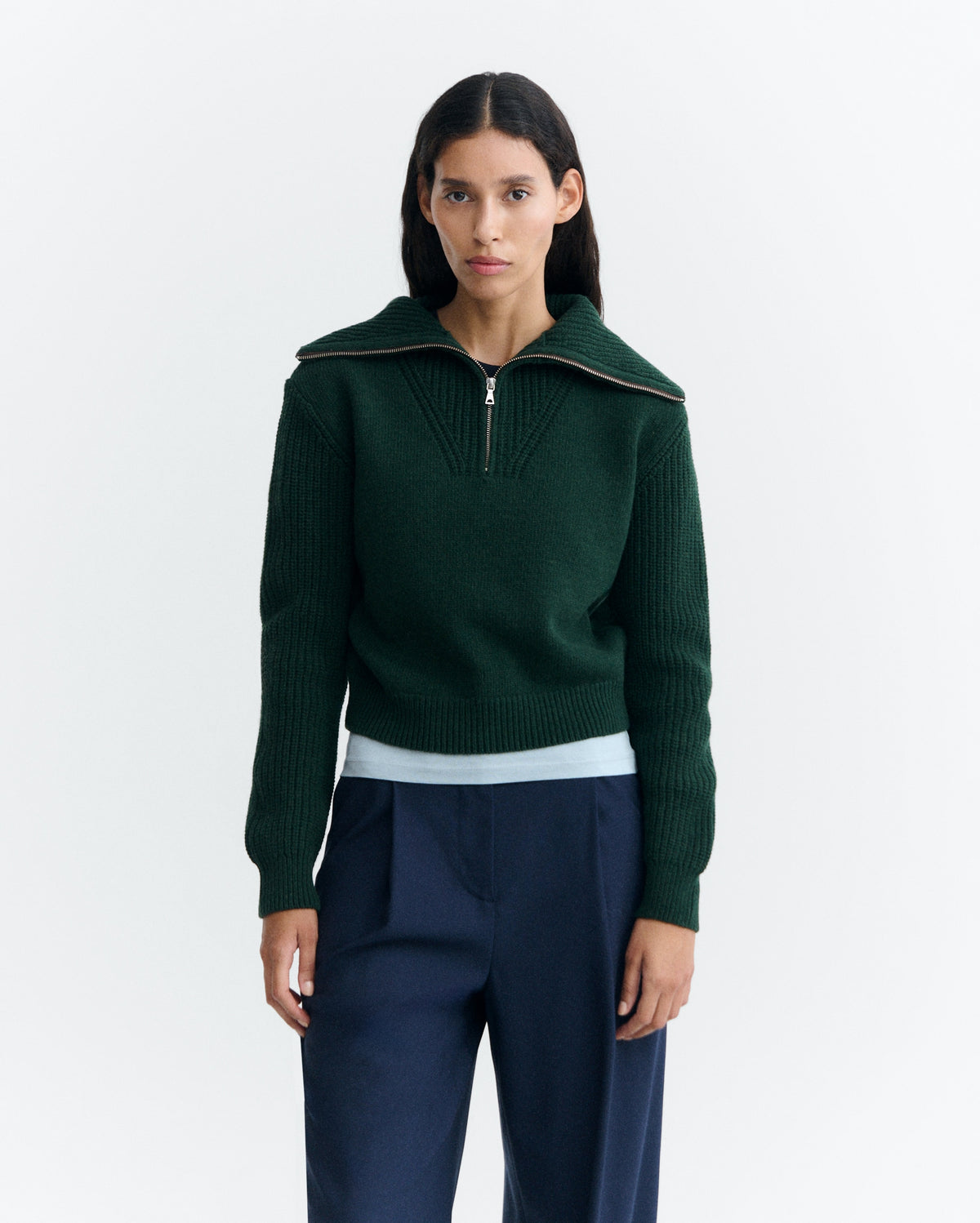 Dunkelgrüner Pullover "Tanit" von Thinking Mu aus Merinowolle mit hohem Kragen, silbernem Reißverschluss und Label am Ausschnitt vor hellem Hintergrund.