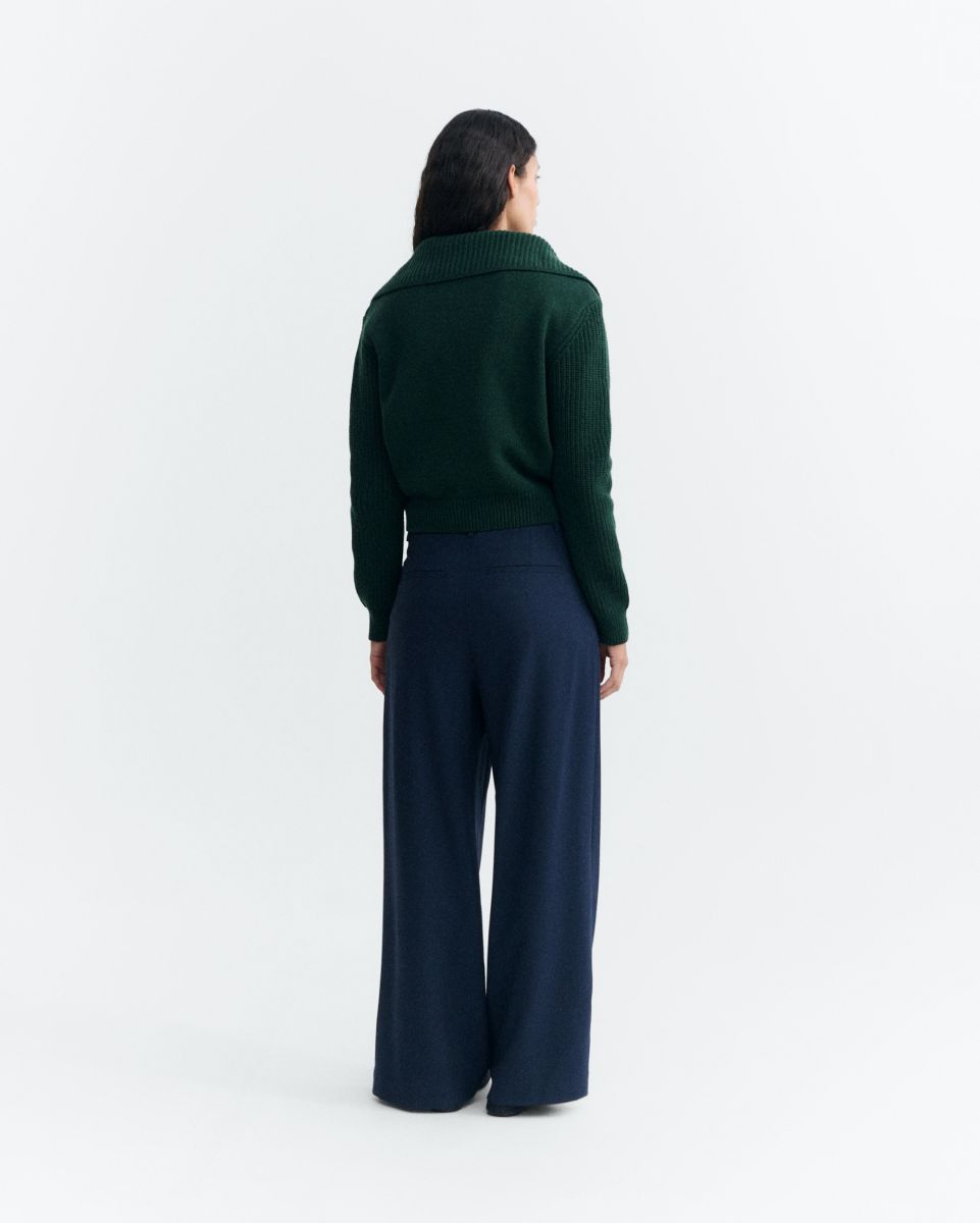 Dunkelgrüner Pullover "Tanit" von Thinking Mu aus Merinowolle mit hohem Kragen, silbernem Reißverschluss und Label am Ausschnitt vor hellem Hintergrund.