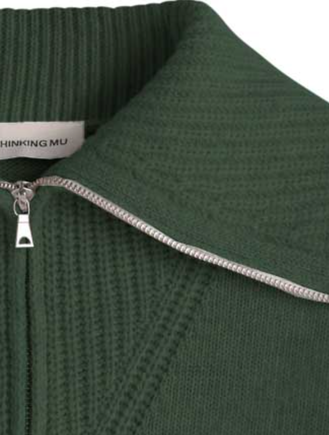 Dunkelgrüner Pullover "Tanit" von Thinking Mu aus Merinowolle mit hohem Kragen, silbernem Reißverschluss und Label am Ausschnitt vor hellem Hintergrund.