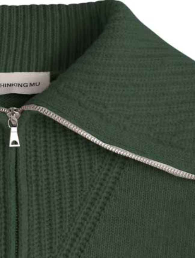 Dunkelgrüner Pullover "Tanit" von Thinking Mu aus Merinowolle mit hohem Kragen, silbernem Reißverschluss und Label am Ausschnitt vor hellem Hintergrund.