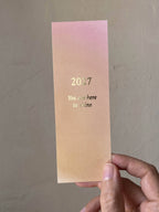 Ein Lesezeichen von Anna Cosma mit rosagelbem Farbverlauf und goldfoliiertem Aufdruck „2027 You are here to shine“ sowie Kalenderübersicht auf der Rückseite.