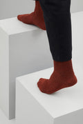 Ein Paar dicke, rustikal gestrickte Socken in Rostrot mit einem gerippten Bündchen. Die Bündchen haben ein Muster aus rostroten und braunen Streifen. Die Socken sind seitlich liegend vor einem weißen Hintergrund dargestellt.