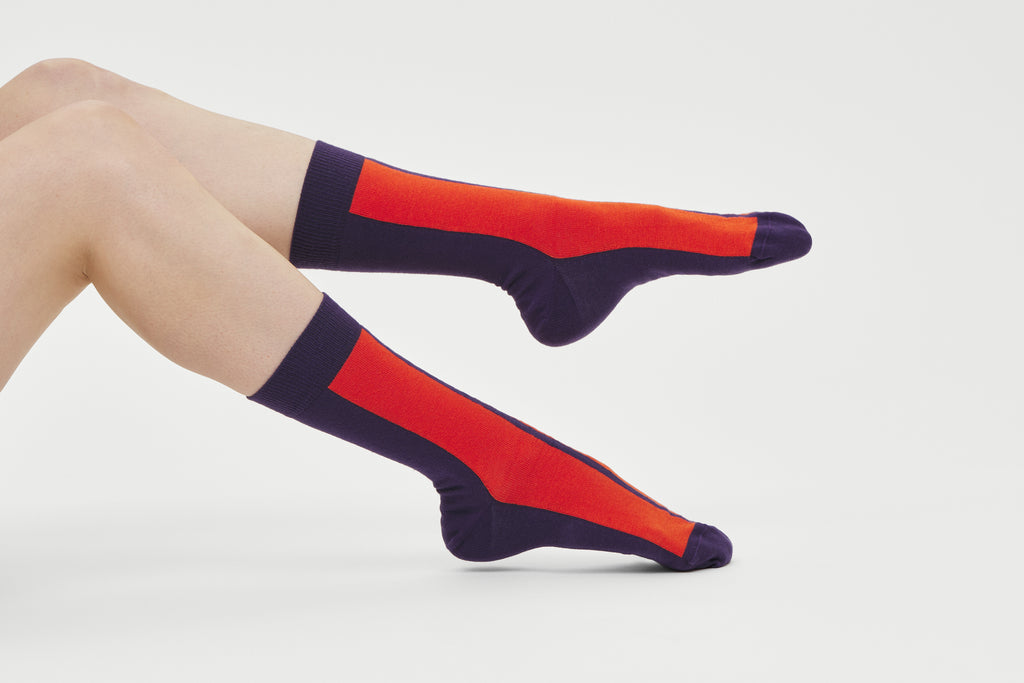 Ein Paar dunkelviolette Socken mit einem auffälligen orangen vertikalen Streifen auf der Vorderseite und dem Markenlogo auf der Fußsohle vor hellem Hintergrund.   