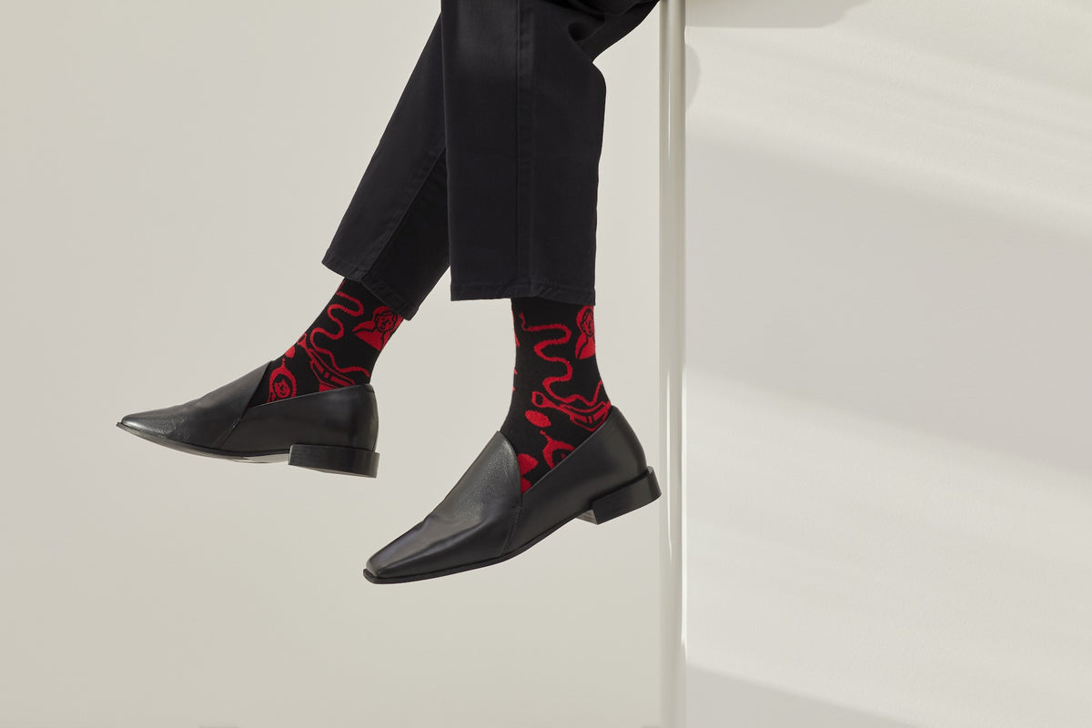 Ein Paar schwarze Socken mit rotem Bund, roter Ferse und Zehenpartie sowie grafischen, roten Figuren- und Tiermotiven auf hellem Hintergrund.