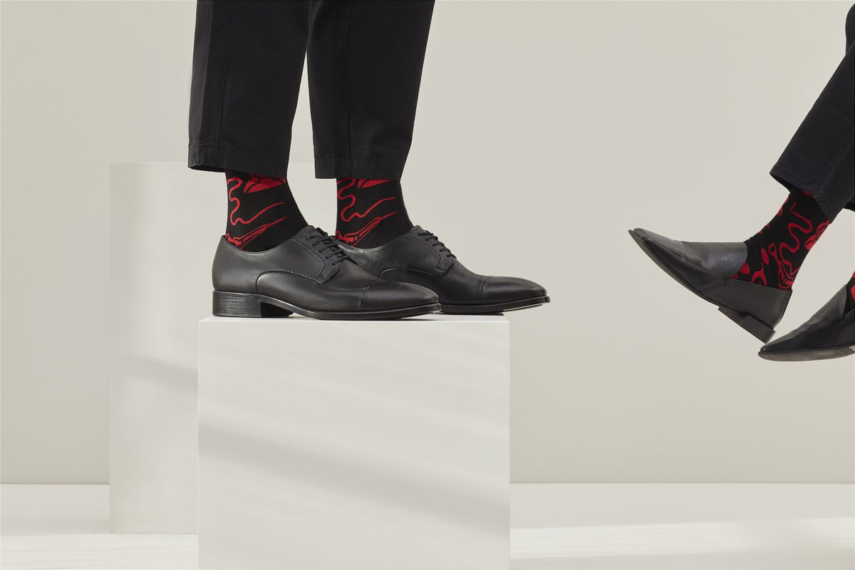 Ein Paar schwarze Socken mit rotem Bund, roter Ferse und Zehenpartie sowie grafischen, roten Figuren- und Tiermotiven auf hellem Hintergrund. koblenz baumwolle