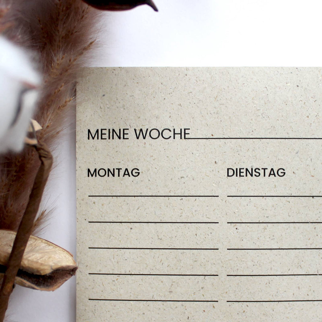 Ein Wochenplaner aus beigem Graspapier mit schwarzen Kornblumen-Illustrationen, Übersicht von Montag bis Sonntag und schwarzer Büroklammer auf weißem Untergrund.