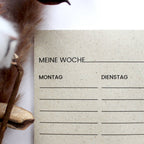 Ein Wochenplaner aus beigem Graspapier mit schwarzen Kornblumen-Illustrationen, Übersicht von Montag bis Sonntag und schwarzer Büroklammer auf weißem Untergrund.