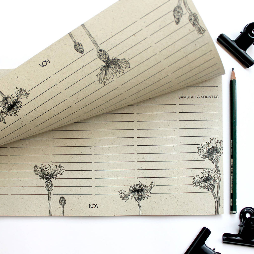 Ein Wochenplaner aus beigem Graspapier mit schwarzen Kornblumen-Illustrationen, Übersicht von Montag bis Sonntag und schwarzer Büroklammer auf weißem Untergrund. koblenz