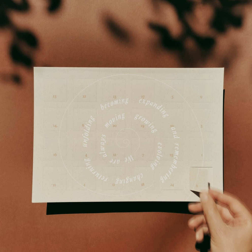 Ein cremefarbener DIN A4 Adventskalender von Anna Cosma mit typografischem Spiral-Motiv und 24 Türchen für achtsame Reflexionsfragen.