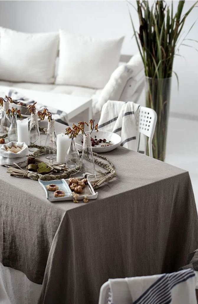 Ein elegant gedeckter Esstisch mit der naturfarbenen (Natural) Epic Linen Tischdecke, dekoriert mit weißen Kerzen, kleinen Gläsern mit Blumen und feinem Gebäck.