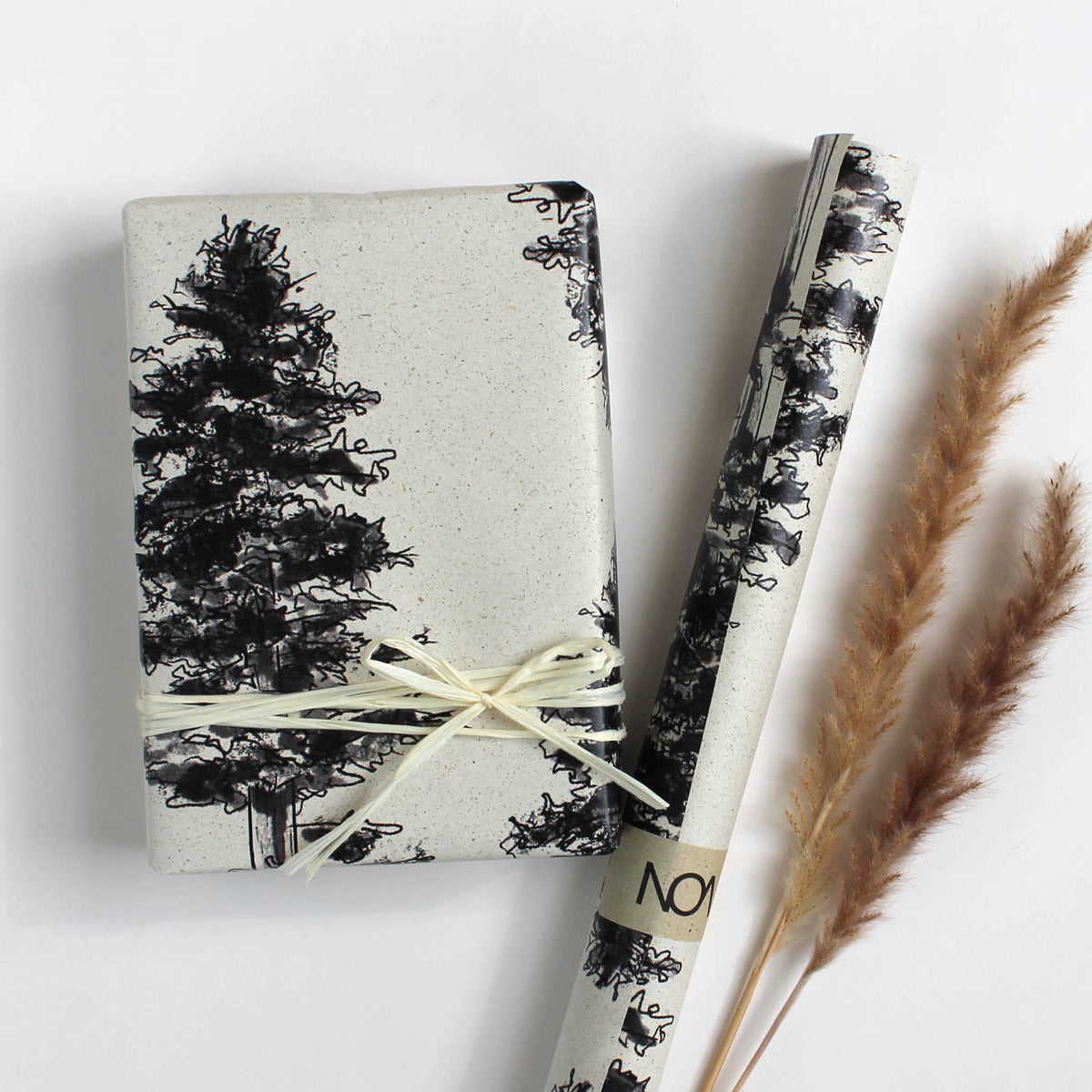 Ein verpacktes Geschenk mit Bastschlaufe und eine Rolle Geschenkpapier aus beigem Graspapier mit gedruckten schwarzen Tannenbäumen.