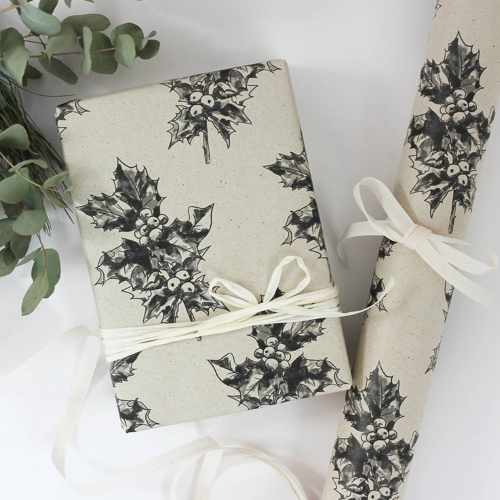 Ein verpacktes Geschenk mit hellem Bastband und aufgerolltes Geschenkpapier aus beigem Graspapier mit handgezeichnetem schwarzen Stechpalmen-Muster.