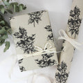 Ein verpacktes Geschenk mit hellem Bastband und aufgerolltes Geschenkpapier aus beigem Graspapier mit handgezeichnetem schwarzen Stechpalmen-Muster.