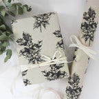 Ein verpacktes Geschenk mit hellem Bastband und aufgerolltes Geschenkpapier aus beigem Graspapier mit handgezeichnetem schwarzen Stechpalmen-Muster.