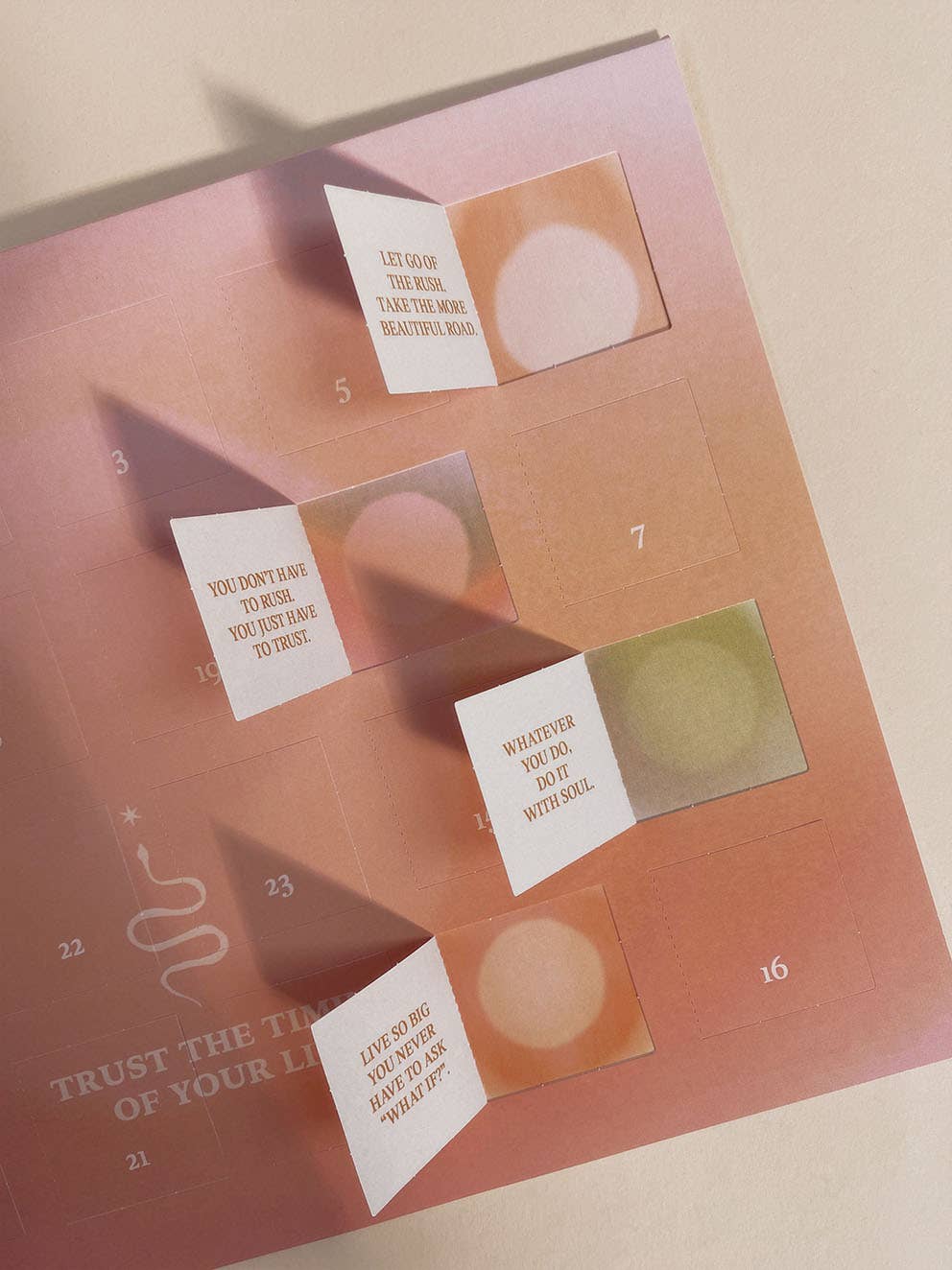 Ein warmtöniger DIN A4 Adventskalender von Anna Cosma mit dem Aufdruck „Trust the timing of your life“, Schlangen-Symbolik und geöffneten Türchen mit Affirmationen. koblenz altstadt trust concept store
