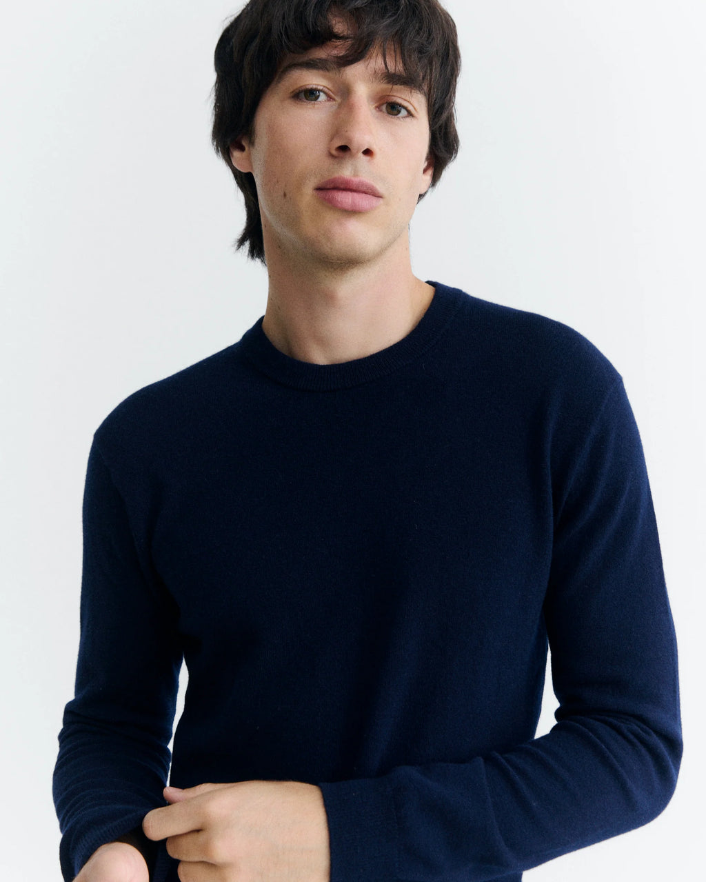  Eine Person mit dunklem Haar trägt einen marineblauen Wollpullover mit Rundhalsausschnitt vor hellem Hintergrund.  