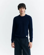  Eine Person mit dunklem Haar trägt einen marineblauen Wollpullover mit Rundhalsausschnitt vor hellem Hintergrund.  