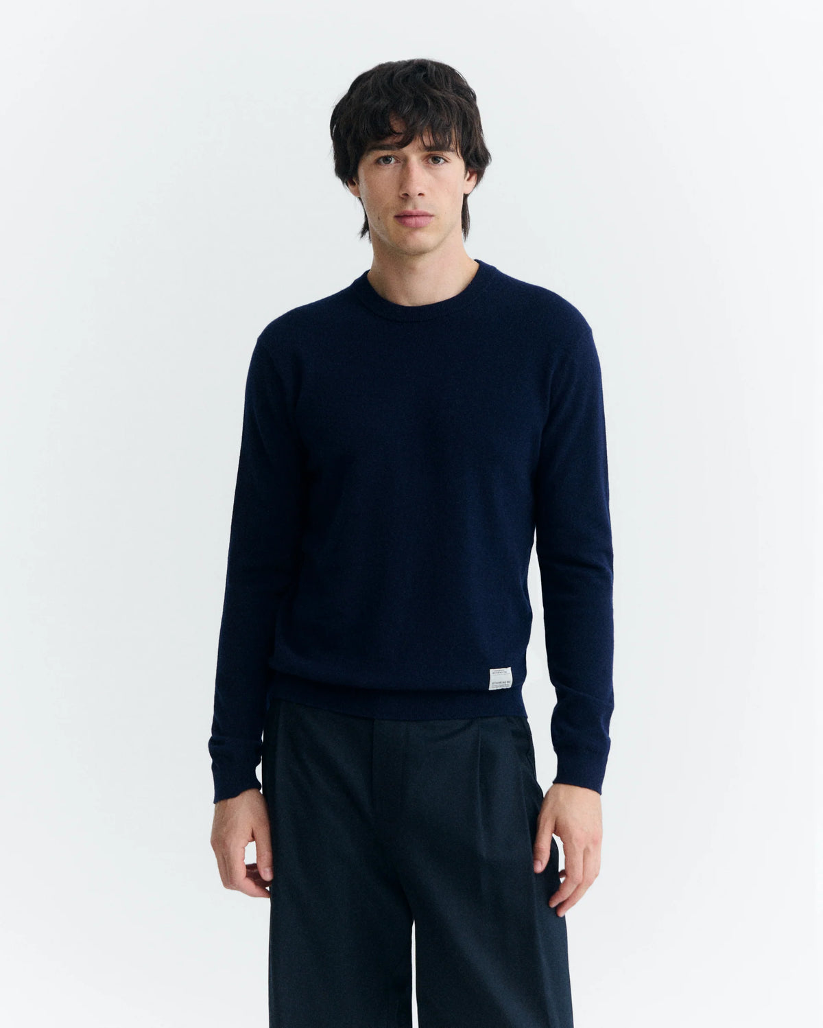  Eine Person mit dunklem Haar trägt einen marineblauen Wollpullover mit Rundhalsausschnitt vor hellem Hintergrund.  
