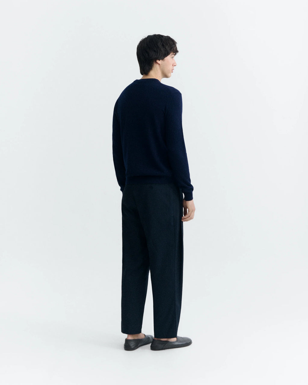  Eine Person mit dunklem Haar trägt einen marineblauen Wollpullover mit Rundhalsausschnitt vor hellem Hintergrund.  