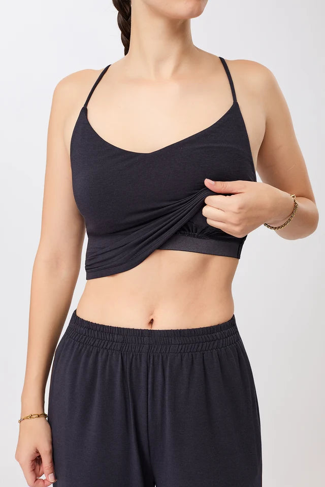 Eine Person trägt ein blaues, ärmelloses Cropped-Top mit V-Ausschnitt und filigranen Trägern zu einer dunklen Hose vor hellem Hintergrund.