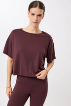 Eine Person trägt ein dunkelrotes, oversized geschnittenes Cropped-T-Shirt mit Rundhalsausschnitt vor hellem Hintergrund