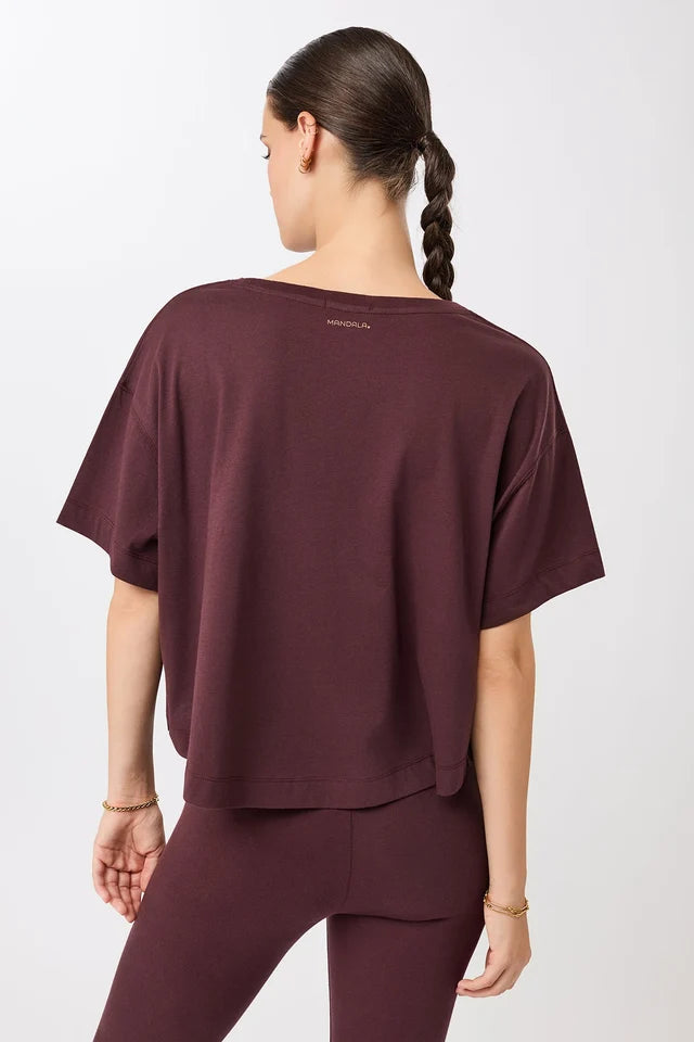 Eine Person trägt ein dunkelrotes, oversized geschnittenes Cropped-T-Shirt mit Rundhalsausschnitt vor hellem Hintergrund