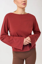 Eine Person trägt ein rotes Langarmshirt mit U-Boot-Ausschnitt und ausgestellten Trompetenärmeln vor hellem Hintergrund.