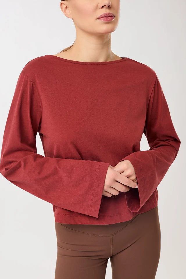 Eine Person trägt ein rotes Langarmshirt mit U-Boot-Ausschnitt und ausgestellten Trompetenärmeln vor hellem Hintergrund.