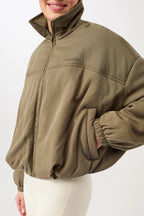 Eine Person trägt eine olivgrüne, sportive Blouson-Jacke mit hohem Stehkragen und Reißverschluss vor hellem Hintergrund