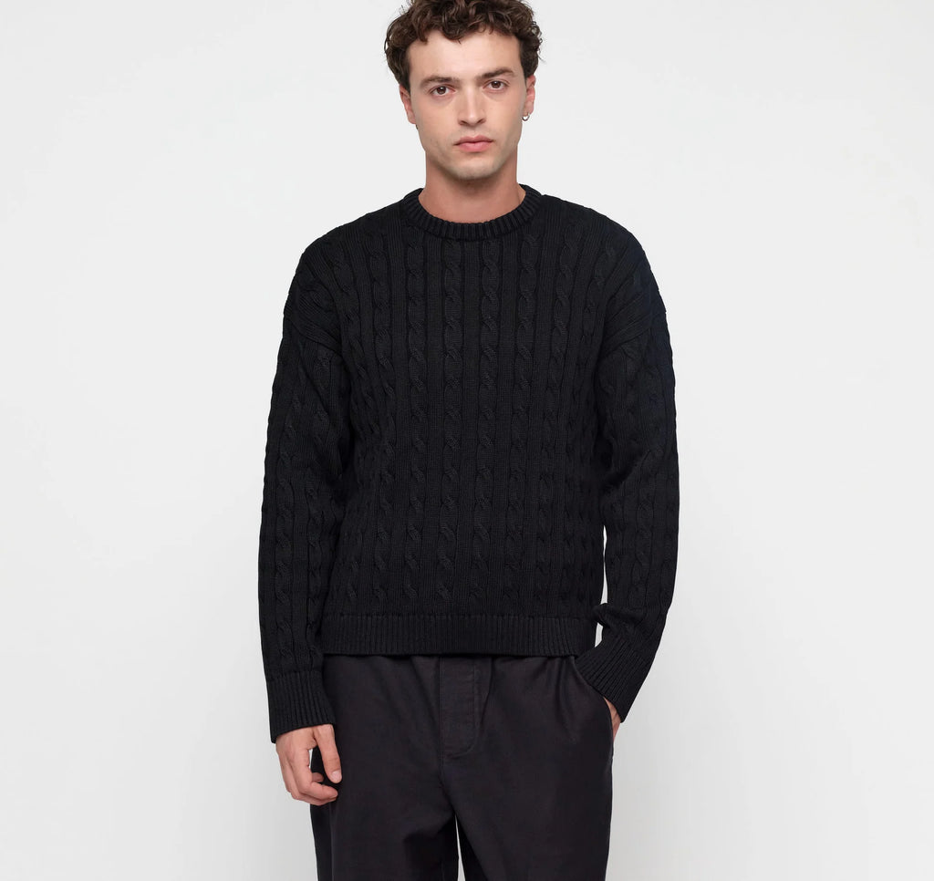 Eine Person trägt einen schwarzen Zopfstrick-Pullover im Boxy Fit mit Rundhalsausschnitt vor hellem Hintergrund. koblenz organic basics