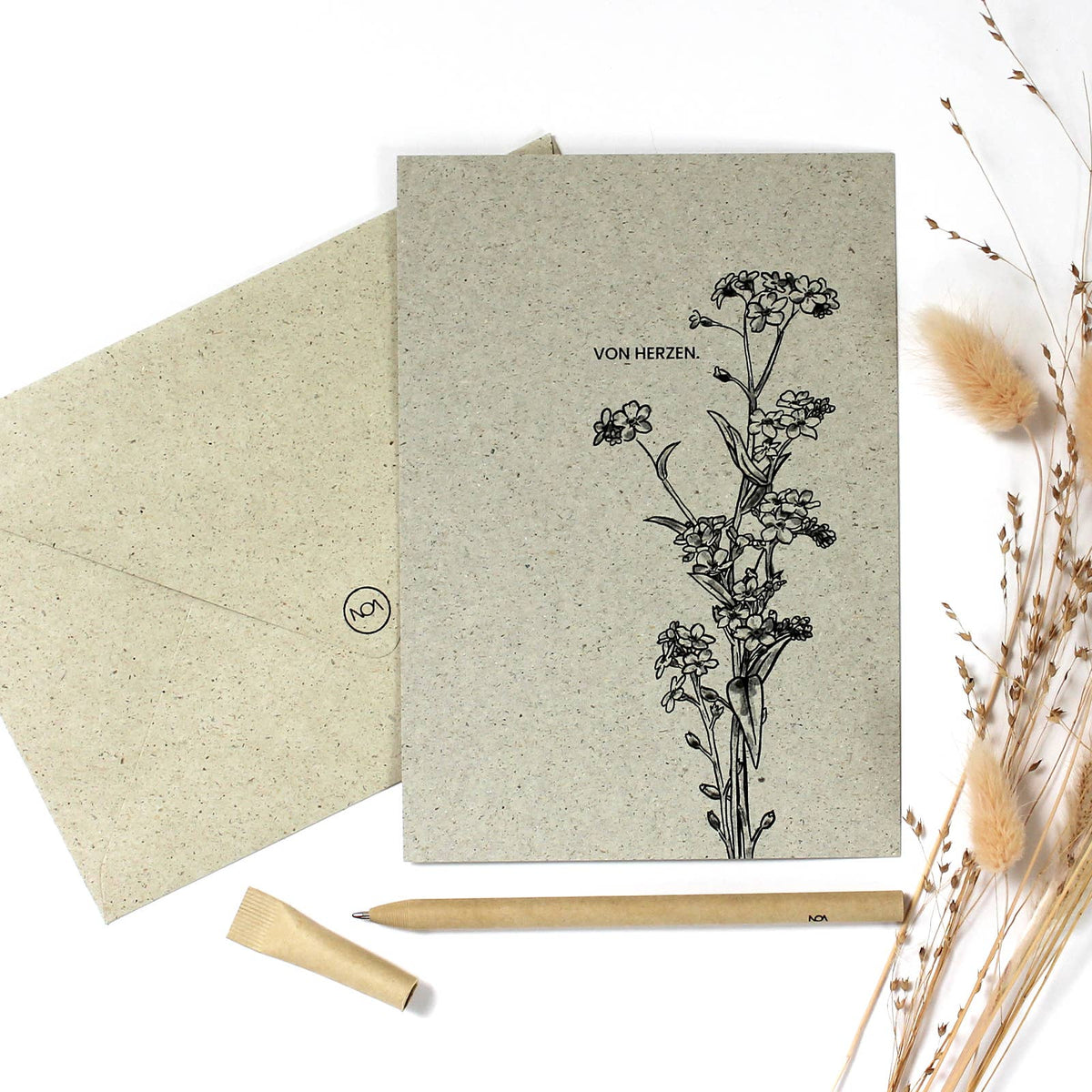 Eine Postkarte aus beigem Graspapier mit schwarzer Handzeichnung einer Vergissmeinnicht-Pflanze und der Aufschrift „Von Herzen.“ neben einem Stift aus Papier.
