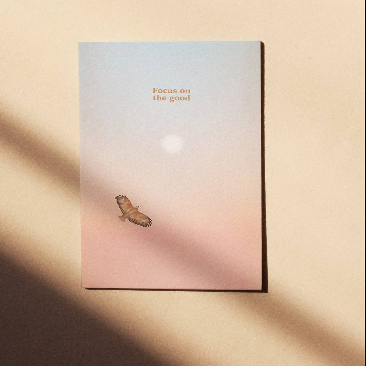 Eine Postkarte mit Rosé-Blau-Farbverlauf, Sonne und fliegendem Greifvogel sowie dem Schriftzug FOCUS ON THE GOOD.