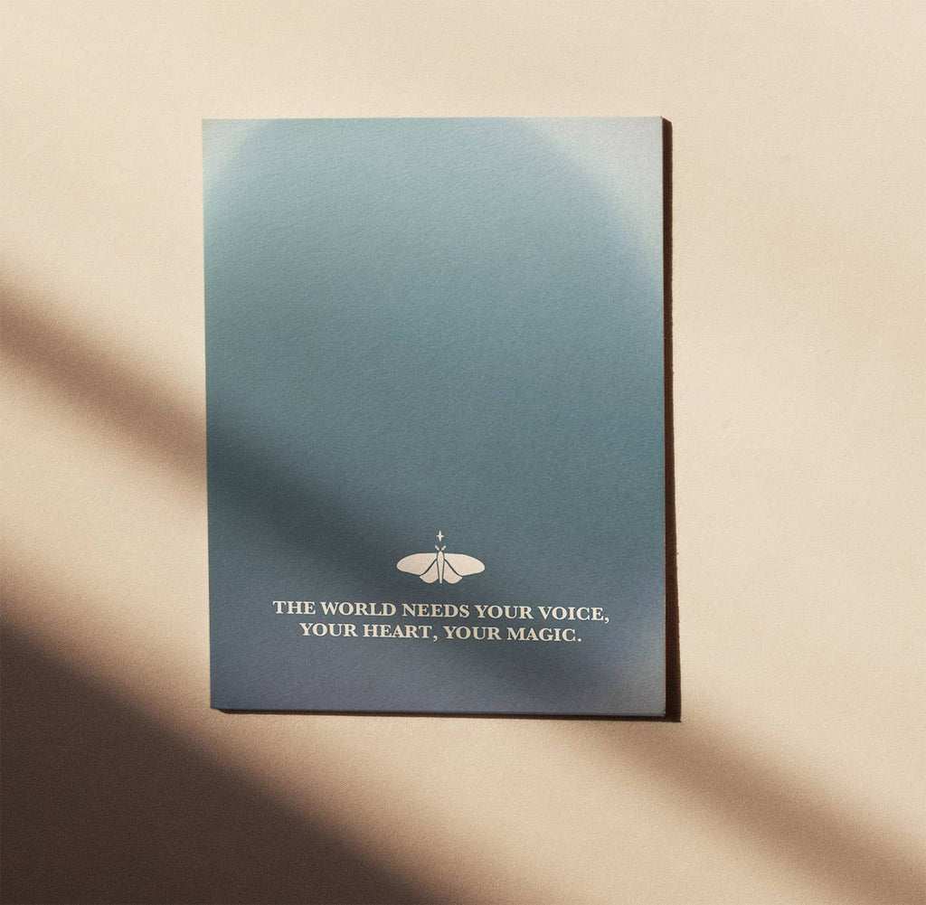 Eine Postkarte von Anna Cosma mit blauem Farbverlauf, Mitten-Mottensymbol und dem englischen Spruch „The world needs your voice, your heart, your magic.“