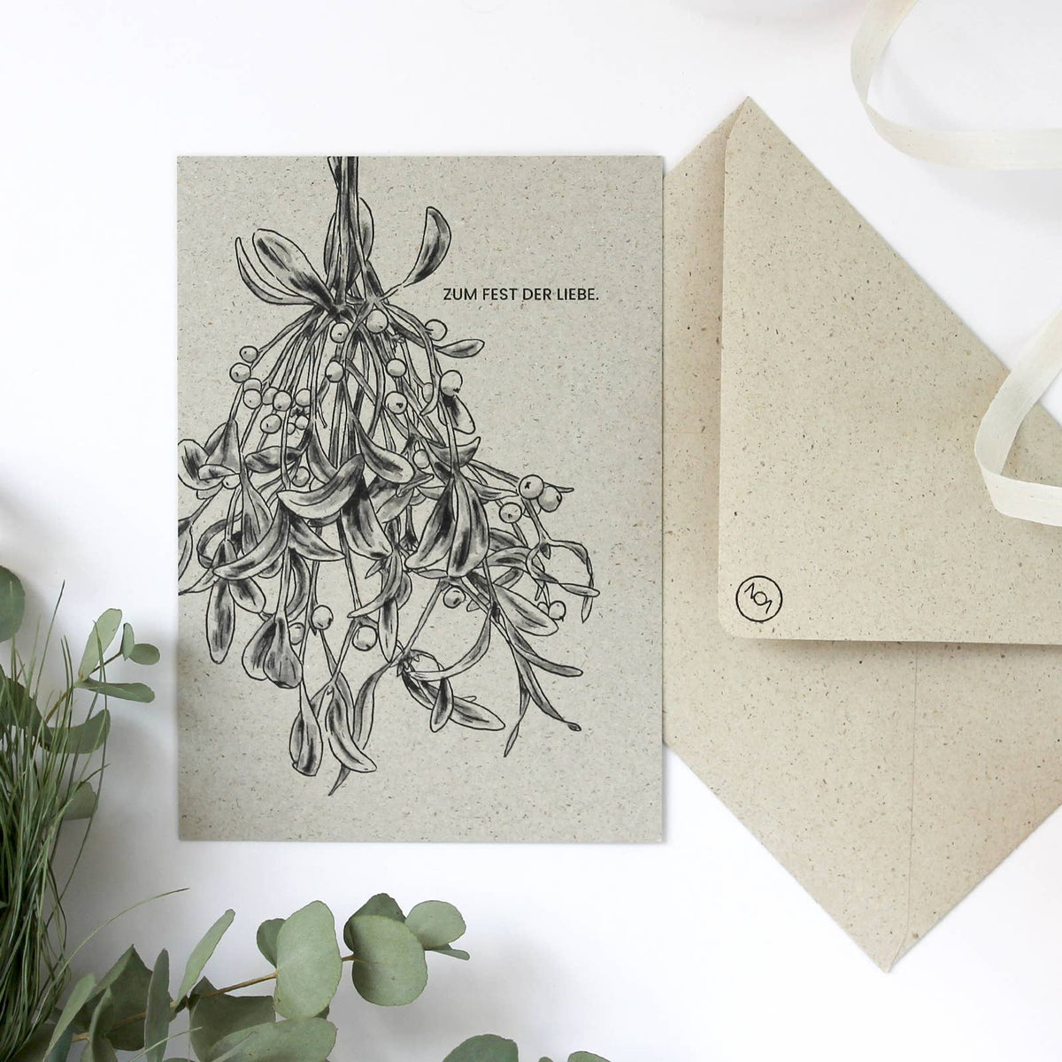 Eine Weihnachtskarte aus naturbelassenem Graspapier mit handgezeichnetem Mistelzweig und dem Text „Zum Fest der Liebe.“ 