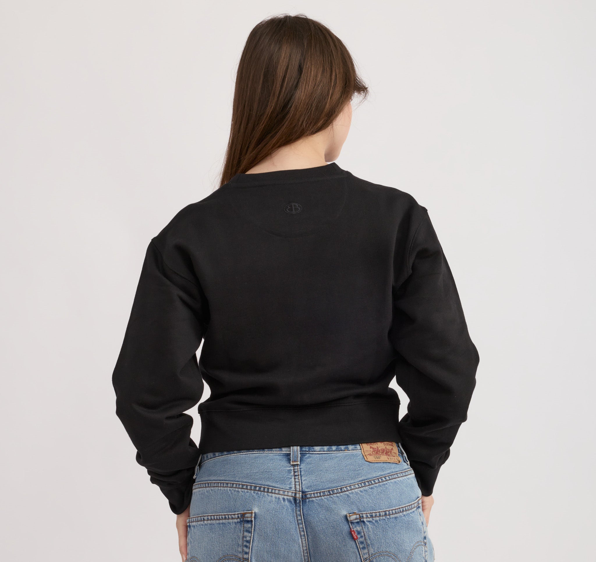 Eine brünette Frau trägt ein schwarzes, kastig geschnittenes Boxy-Sweatshirt mit Rundhalsausschnitt und Blue Jeans vor hellem Hintergrund.
