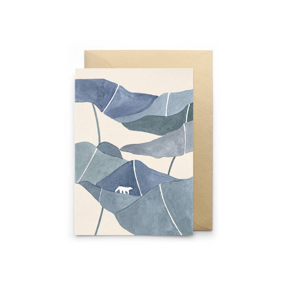 Eine gefaltete Grußkarte mit einer blauen Aquarell-Berglandschaft und kleinem weißen Eisbären samt Kraftpapier-Umschlag vor weißem Hintergrund.