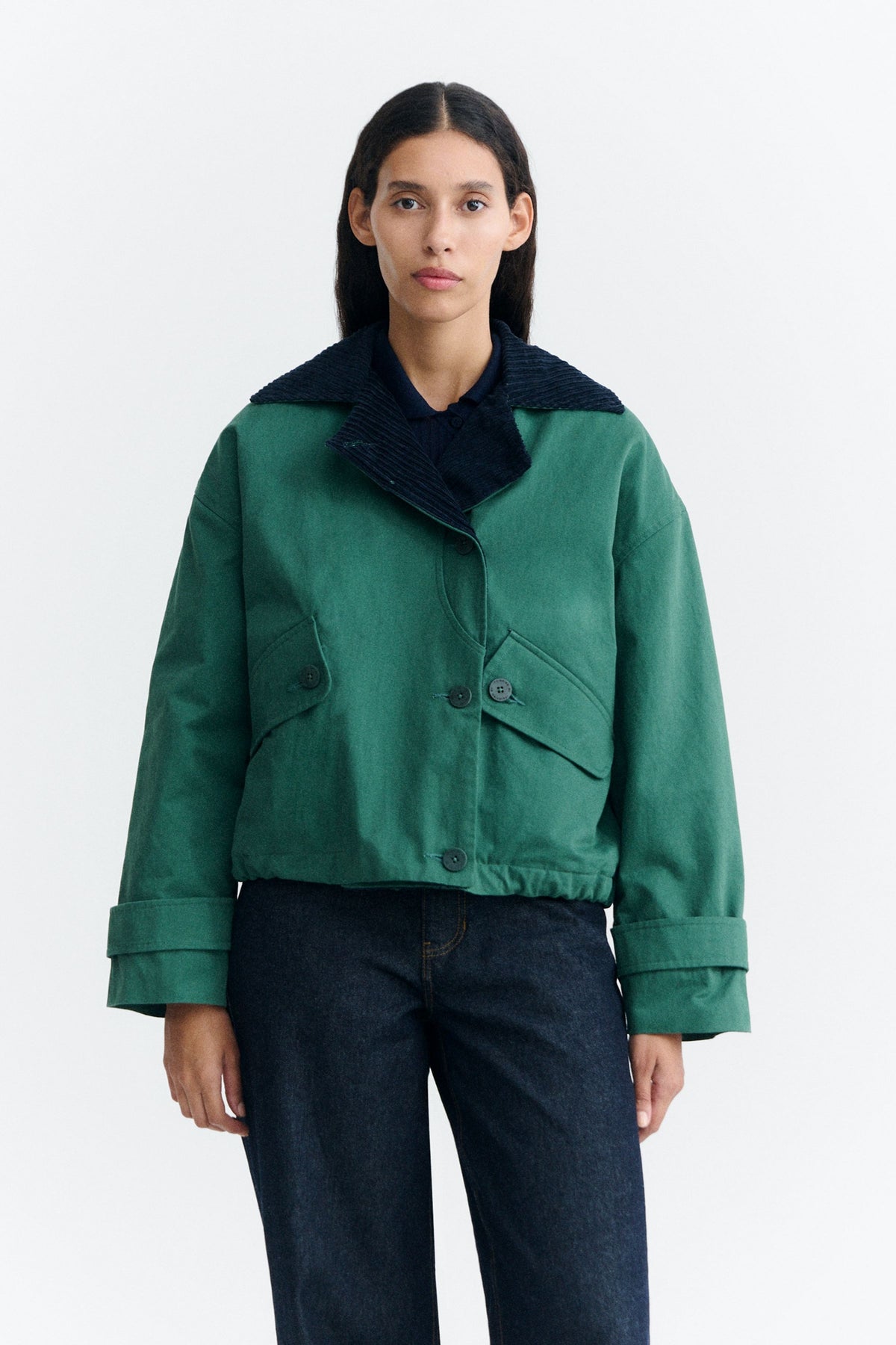 Grüne Jacke "Norma" von Thinking Mu mit dunklem Cord-Kragen, Knöpfen und herabhängenden Bändern am Saum vor hellem Hintergrund.