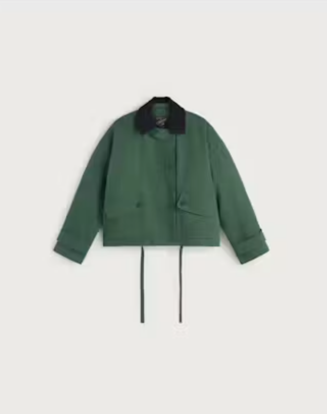 Grüne Jacke "Norma" von Thinking Mu mit dunklem Cord-Kragen, Knöpfen und herabhängenden Bändern am Saum vor hellem Hintergrund.