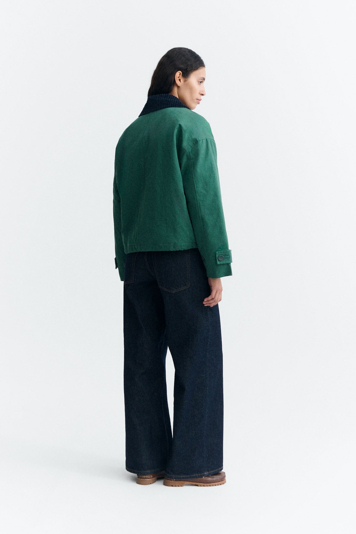 Grüne Jacke "Norma" von Thinking Mu mit dunklem Cord-Kragen, Knöpfen und herabhängenden Bändern am Saum vor hellem Hintergrund.