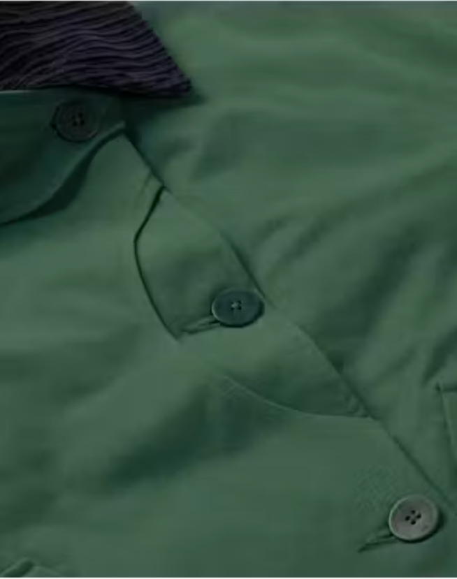 Grüne Jacke "Norma" von Thinking Mu mit dunklem Cord-Kragen, Knöpfen und herabhängenden Bändern am Saum vor hellem Hintergrund.