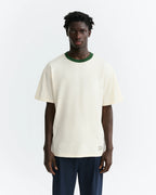 Person von hinten in einem hellen T-Shirt "Aaron" mit grünem Pirineo-Print auf der Rückseite und kontrastfarbenem grünen Rippkragen vor hellem Hintergrund.