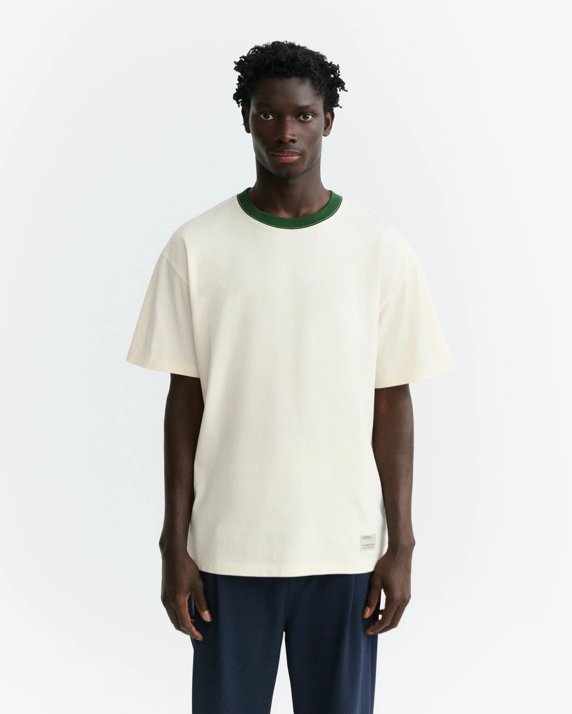 Person von hinten in einem hellen T-Shirt "Aaron" mit grünem Pirineo-Print auf der Rückseite und kontrastfarbenem grünen Rippkragen vor hellem Hintergrund.