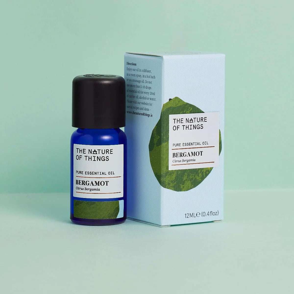 Kleine blaue 12-ml-Flasche des Essential Oil Bergamot von The Nature of Things und dazugehörige hellblaue Umverpackung mit Bergamotte-Illustration vor einem hellgrünen Hintergrund.