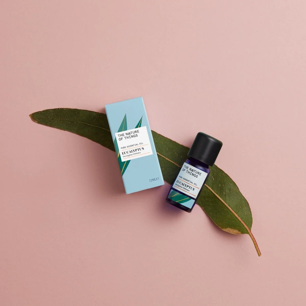 Kleine blaue 12-ml-Flasche des Essential Oil Organic Eucalyptus von The Nature of Things und dazugehörige hellblaue Umverpackung mit Eukalyptus-Illustrationen vor einem beigen Hintergrund.