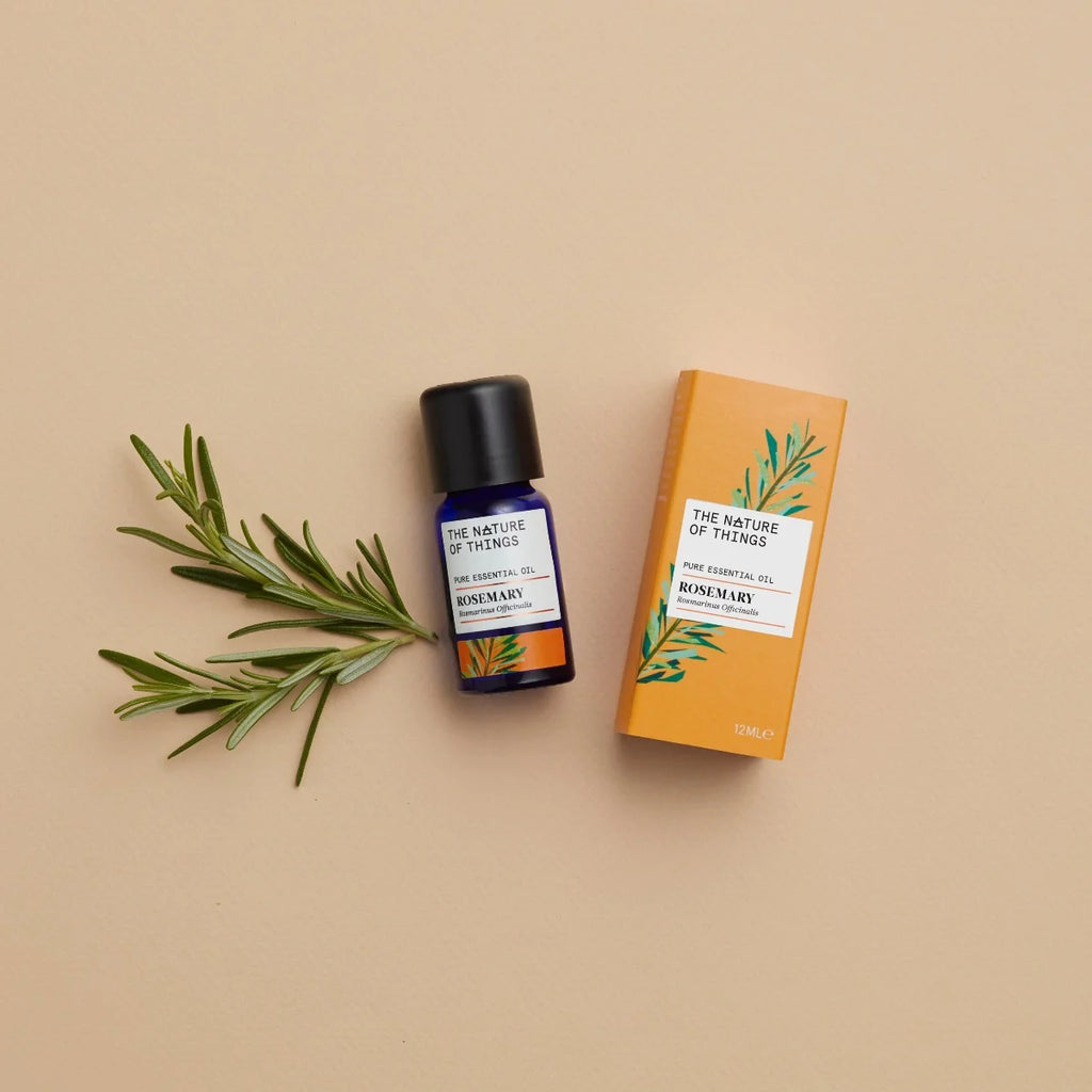 Kleine blaue 12-ml-Flasche des Essential Oil Organic Rosemary von The Nature of Things und dazugehörige orangefarbene Umverpackung mit Rosmarin-Illustrationen vor einem beigen Hintergrund.